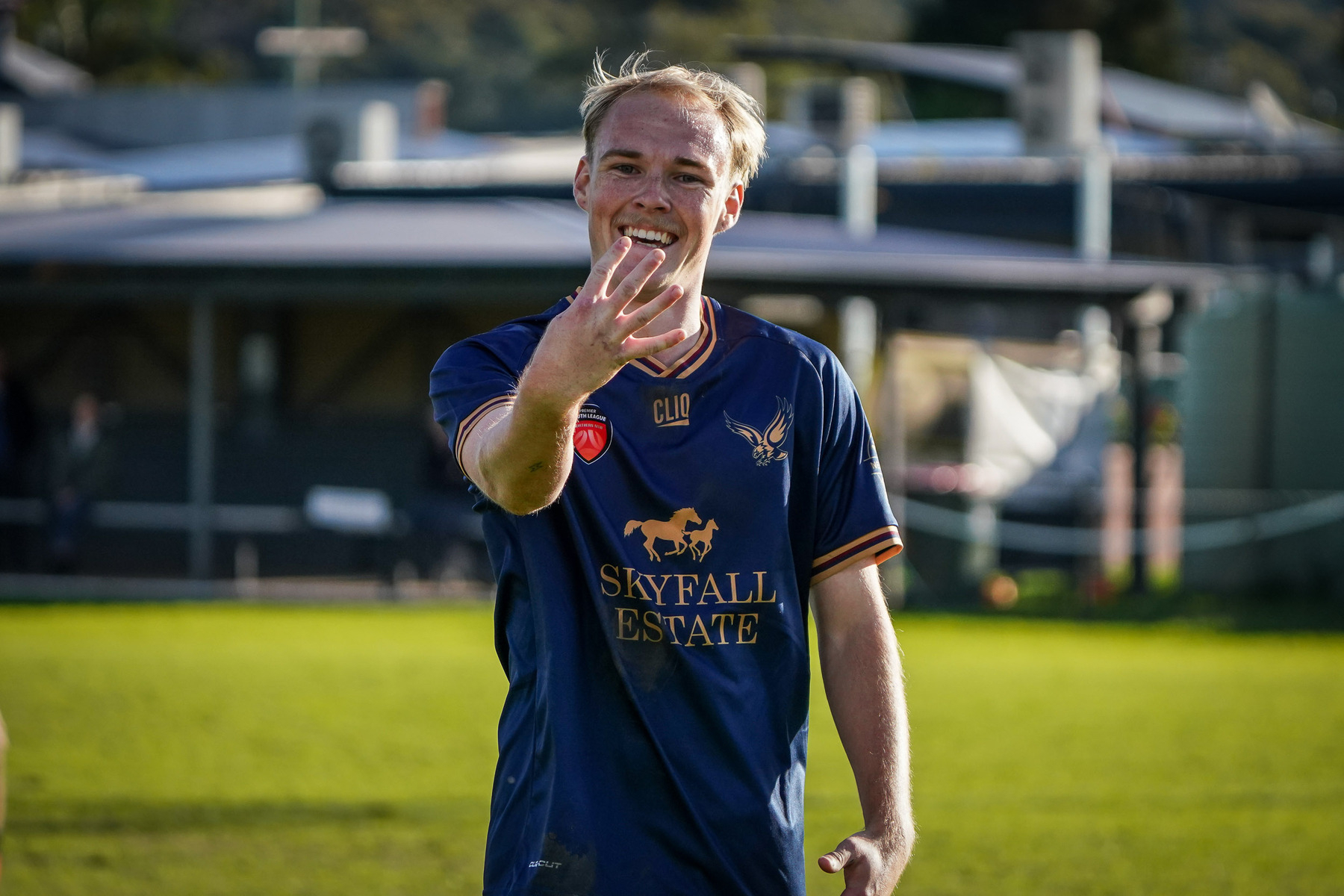 A soccer player in a blue jersey is standing on a field, smiling and holding up four fingers.