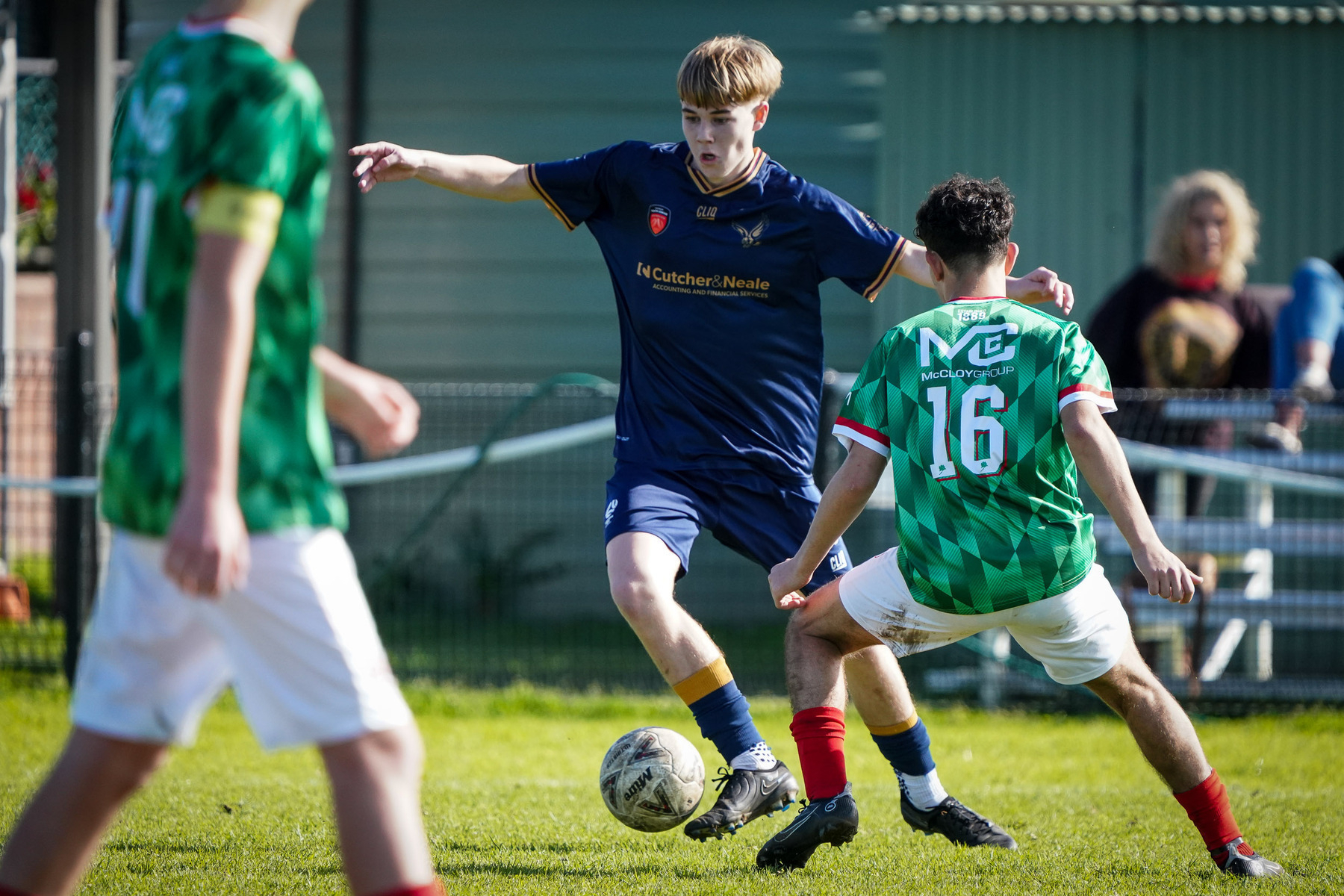 A soccer match is in progress with one player in a navy blue jersey maneuvering the ball while being challenged by another in a green checkered jersey, observed by people in the background.