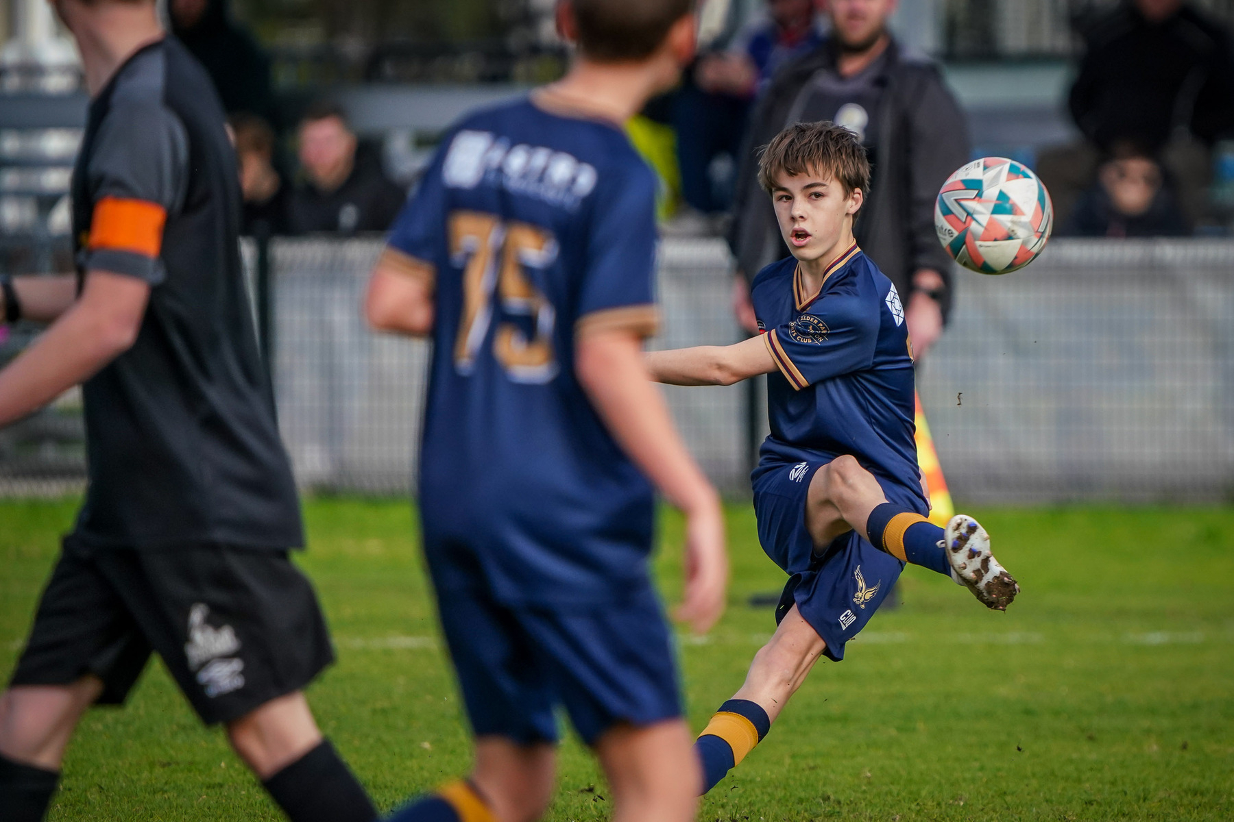 A young soccer player in a blue uniform is mid-kick during a match, with teammates and opponents visible in the background.