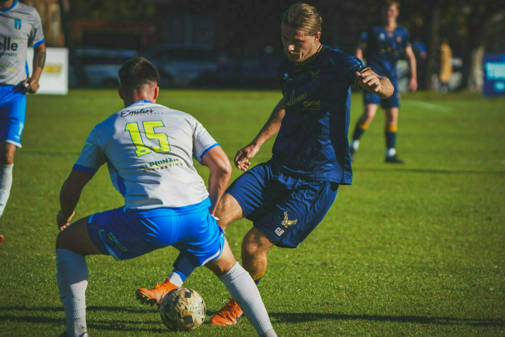 Two soccer players are engaged in a match, one in a white and blue uniform and the other in a dark blue uniform, as they compete for possession of the ball on a grassy field.