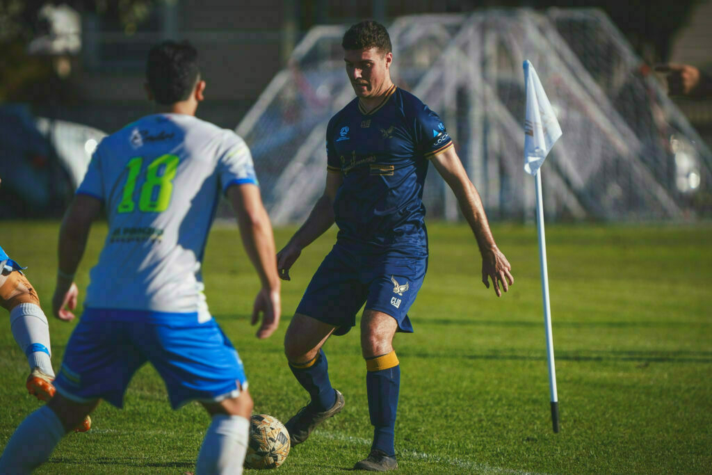 Two soccer players wearing blue compete for the ball near a corner flag on a sunny field.