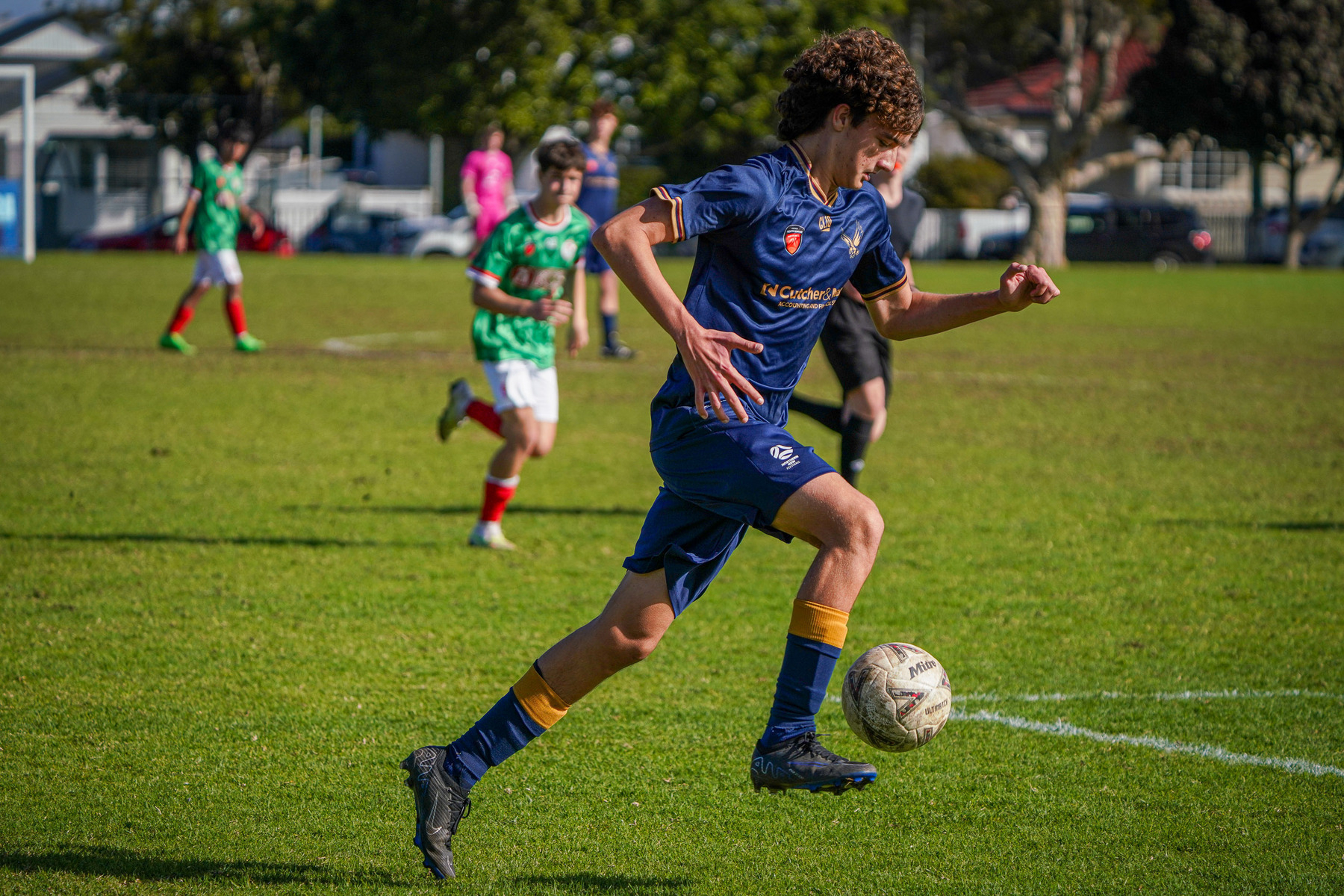 A young soccer player in a blue and yellow uniform is dribbling a ball on a grassy field during a match.