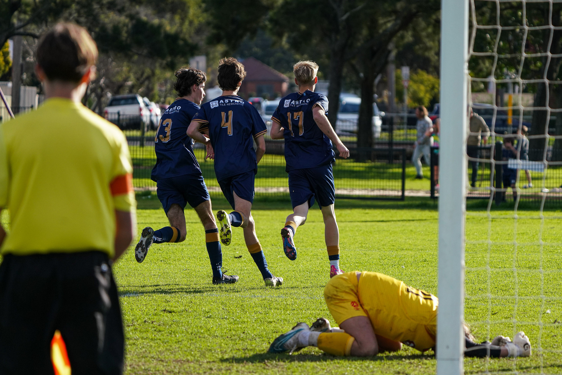 Three soccer players in blue jerseys run in celebration while a goalkeeper in yellow lies on the ground, and a referee looks on.