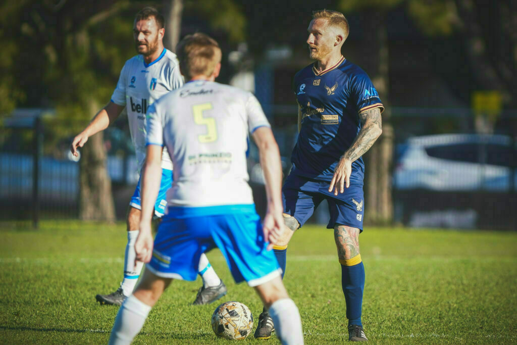 Two soccer players in white and blue uniforms face an opposing player in a dark blue uniform during a match.