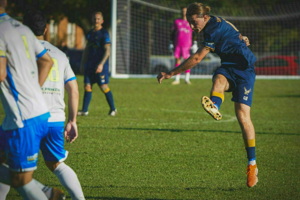 A soccer player in blue is mid-kick during a match on a grass field, with other players and a goal visible in the background.