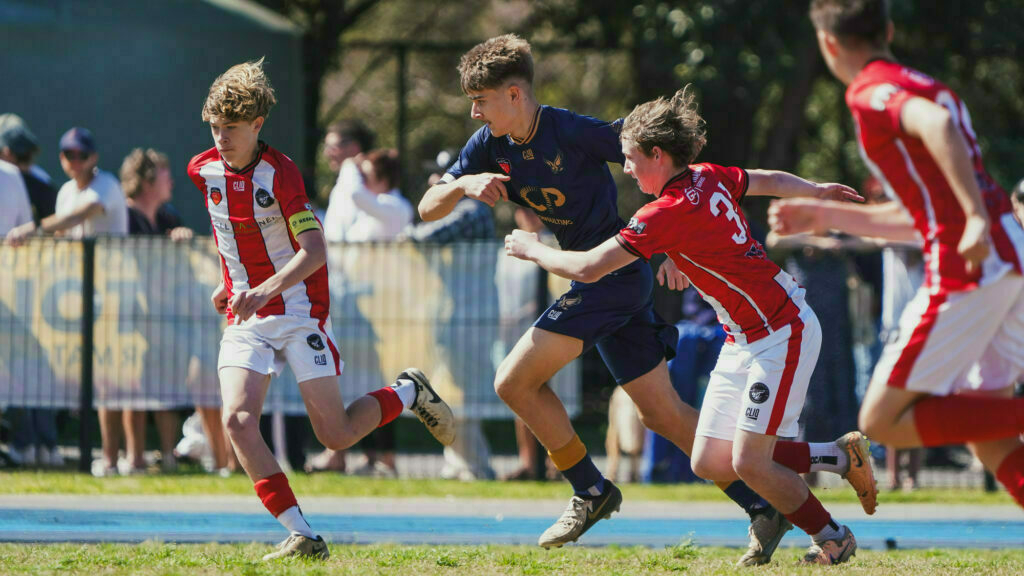 A group of young athletes in red and blue uniforms are actively playing outdoor soccer, with spectators watching from the sidelines.