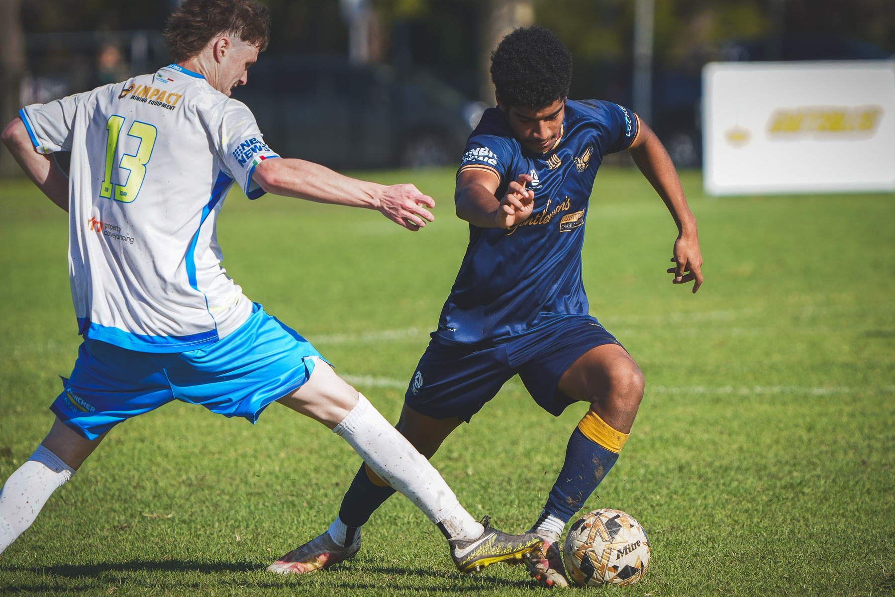 Two soccer players are engaged in a match, with one attempting to maneuver the ball past the other on a grassy field.