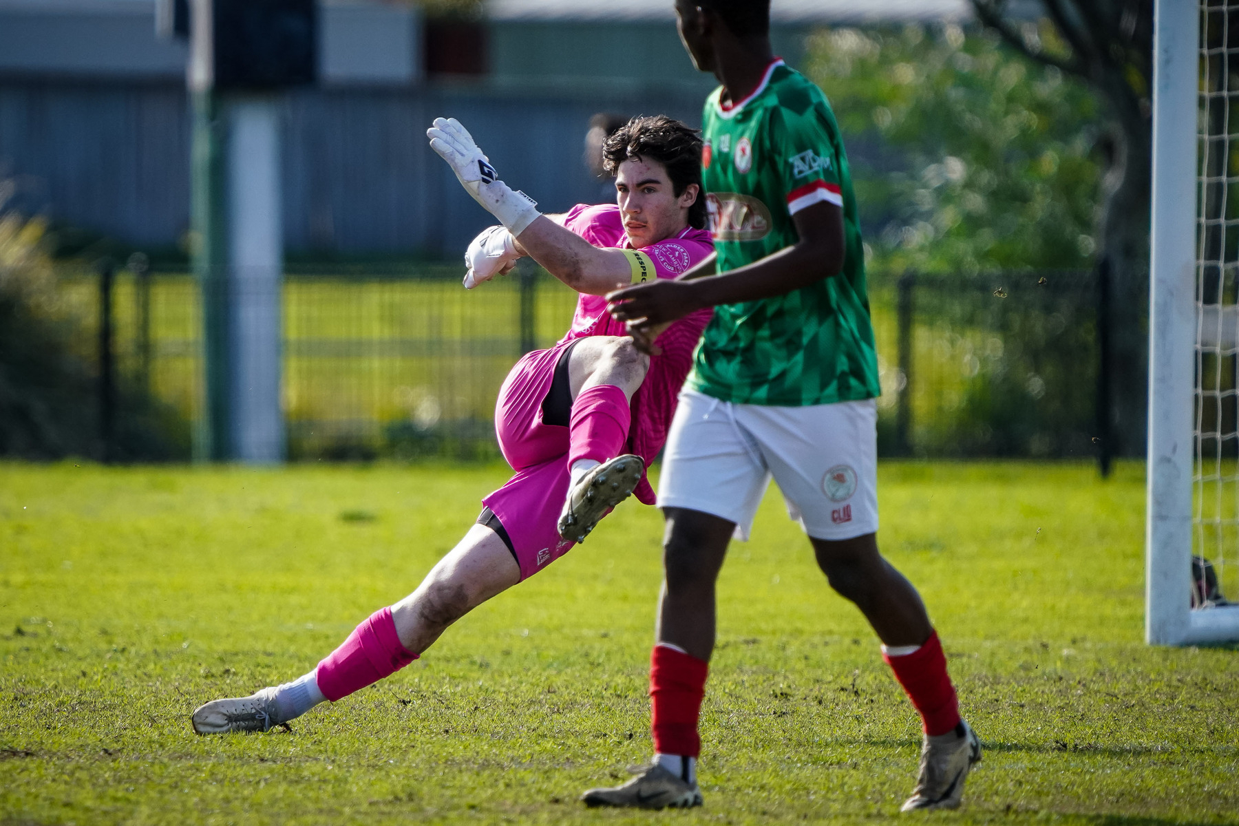 A goalkeeper in a pink uniform is making a dynamic save during a soccer match while an opposing player looks on.