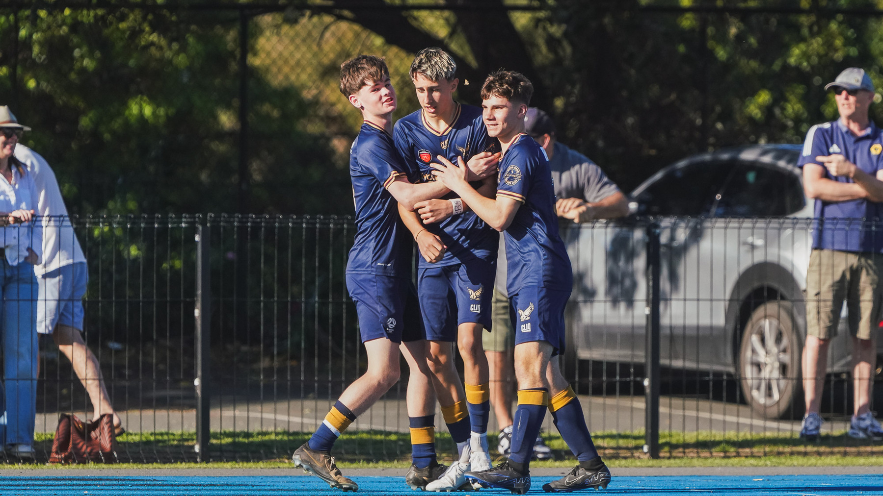 Three young soccer players in matching uniforms are celebrating together near the field.