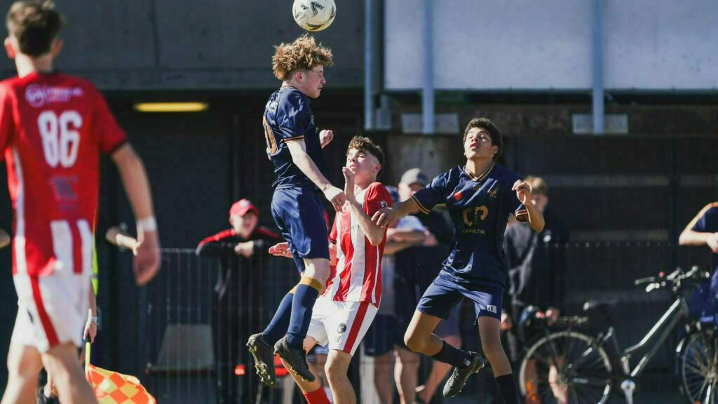 Three soccer players compete for a header during a match, with surrounding players and spectators in the background.