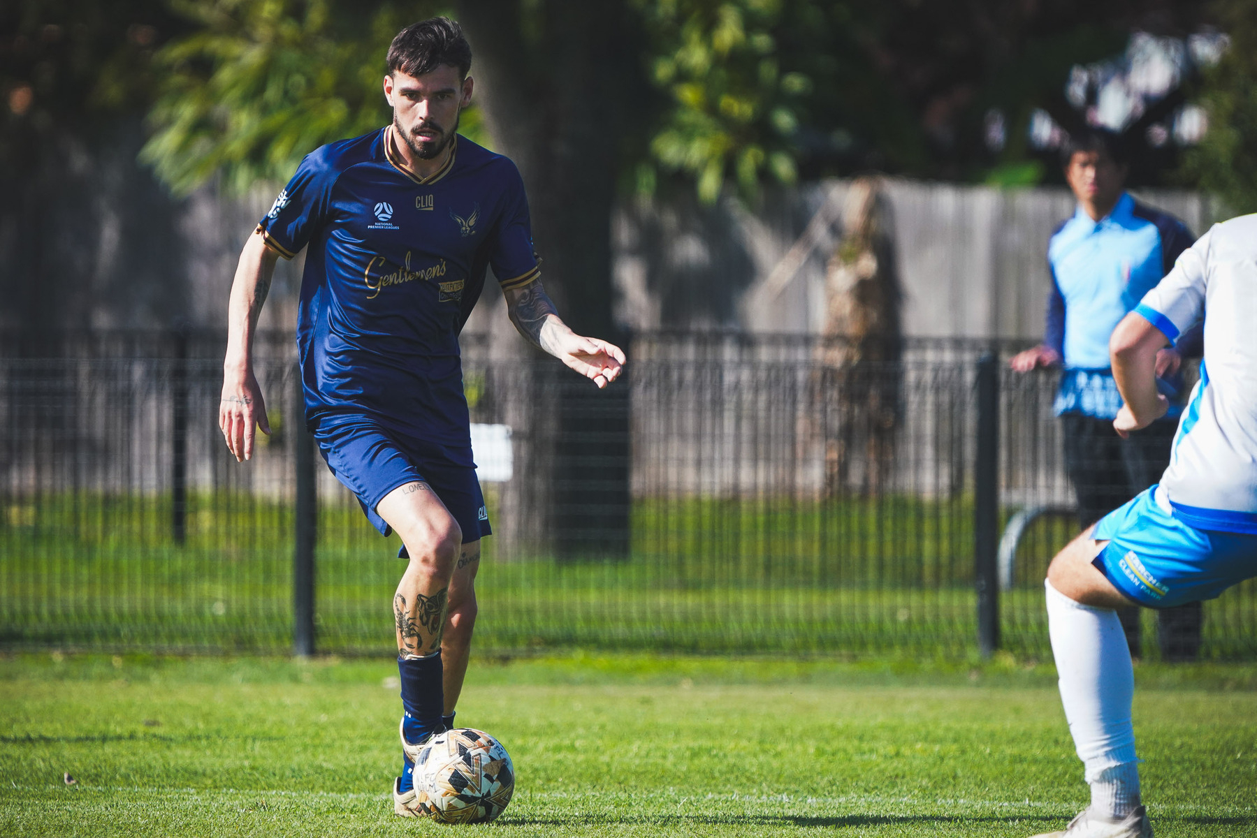 A soccer player in a blue uniform is controlling the ball while being approached by an opponent on a grassy field.