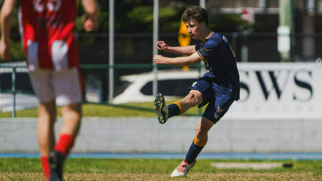 A young soccer player in a blue and yellow uniform makes a powerful kick on the field, while another player in a red and white uniform stands nearby.