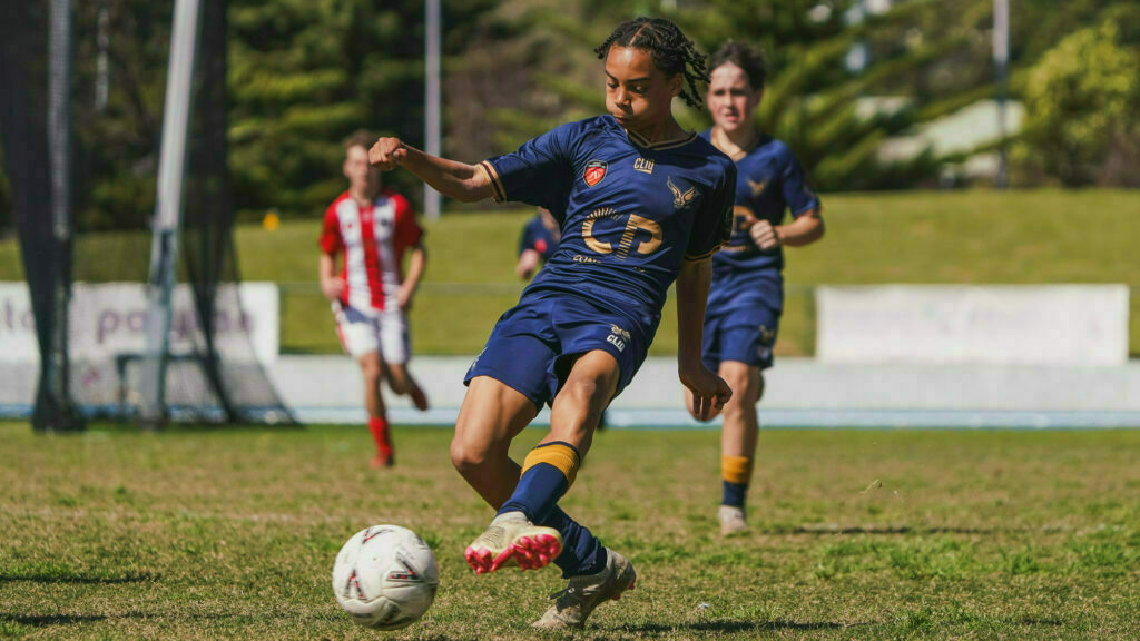 A young soccer player in a navy blue uniform is about to kick a ball during a match on a grassy field, with other players in the background.