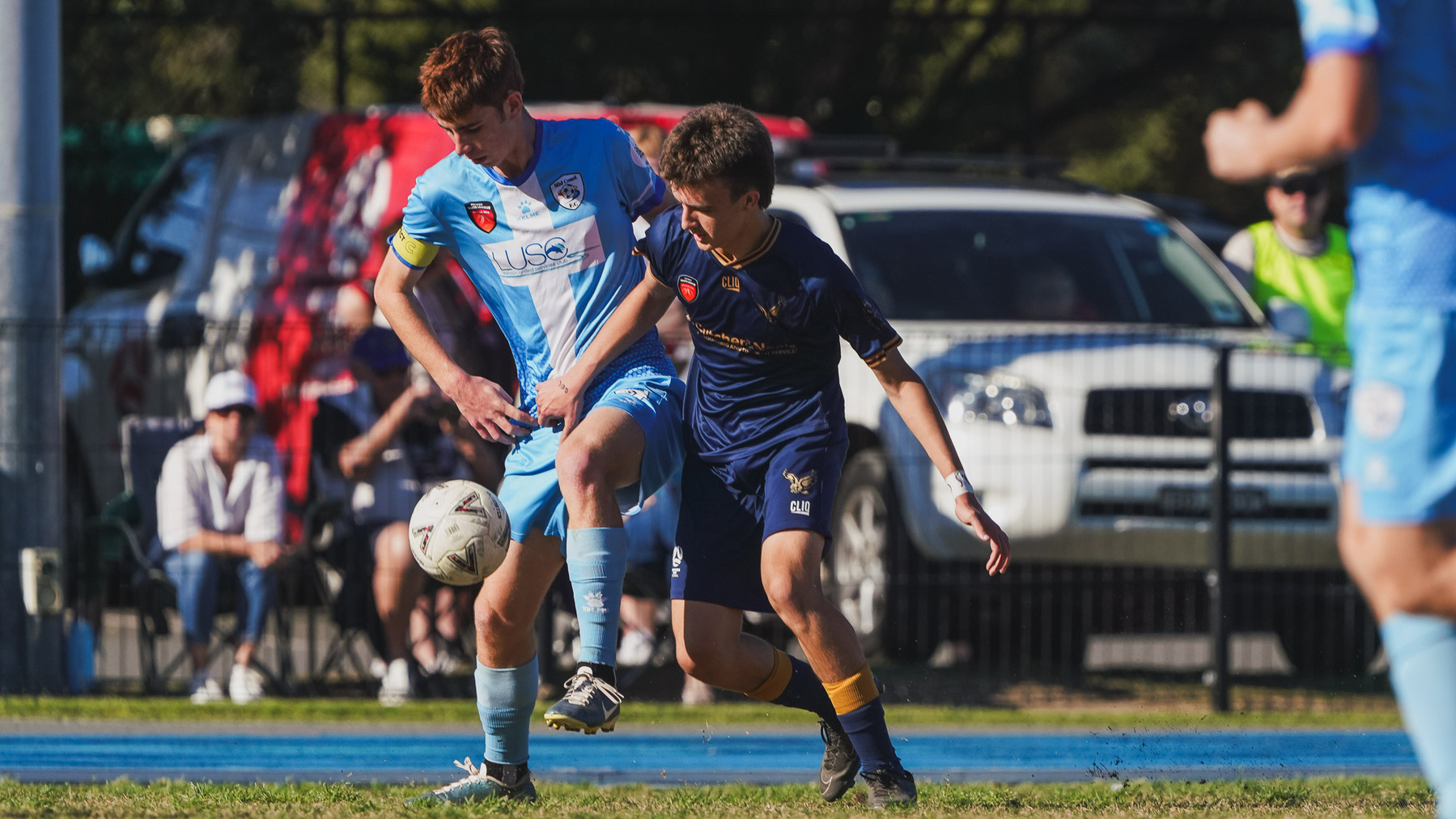 Two soccer players in different team uniforms are competing for the ball during a match, with spectators and parked cars in the background.