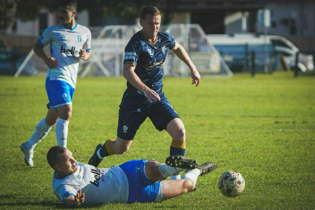 Two soccer players in blue and white uniforms are actively competing for the ball on a grassy field, with one player on the ground.