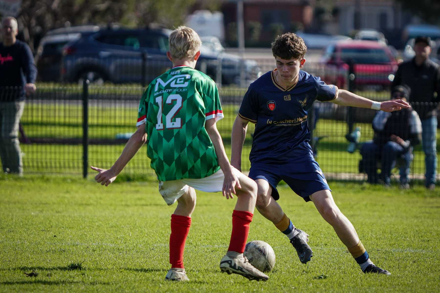 Two young soccer players are engaged in a match, one wearing a green jersey with number 12, as spectators watch from the sidelines.