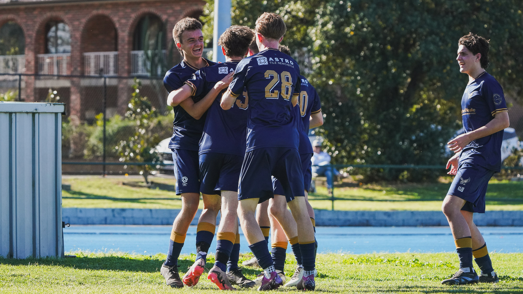 A group of young soccer players in navy uniforms are celebrating on a grassy field.