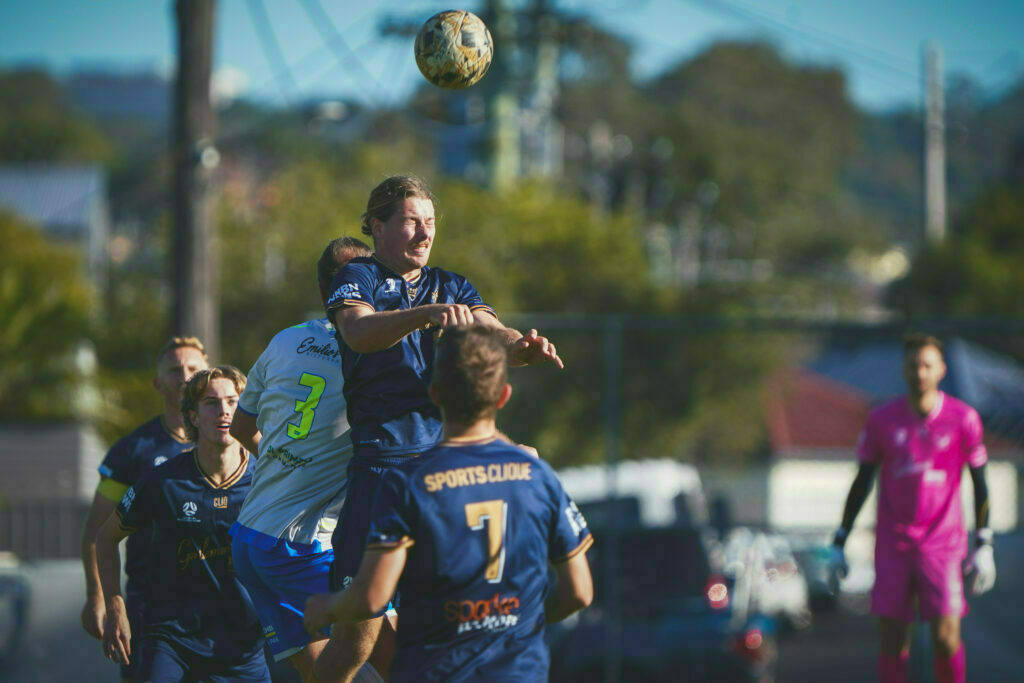 Two soccer players are jumping to head the ball during a match, with other players and a referee visible in the background.