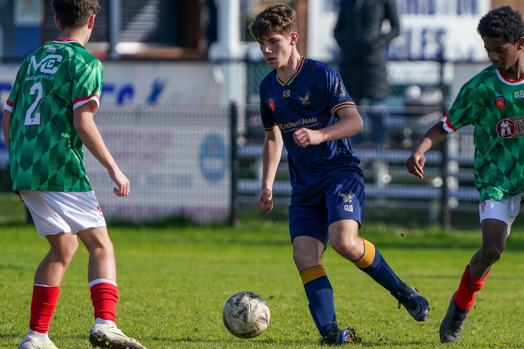 A soccer player in a blue uniform is dribbling the ball while being pursued by two opposing players in green uniforms.