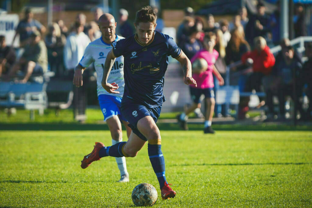 A soccer player in a dark blue uniform is dribbling a ball on a grassy field with an opponent nearby and spectators in the background.