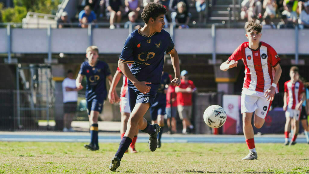 A young soccer player in a navy uniform dribbles the ball on a grassy field during a match, with other players and spectators in the background.