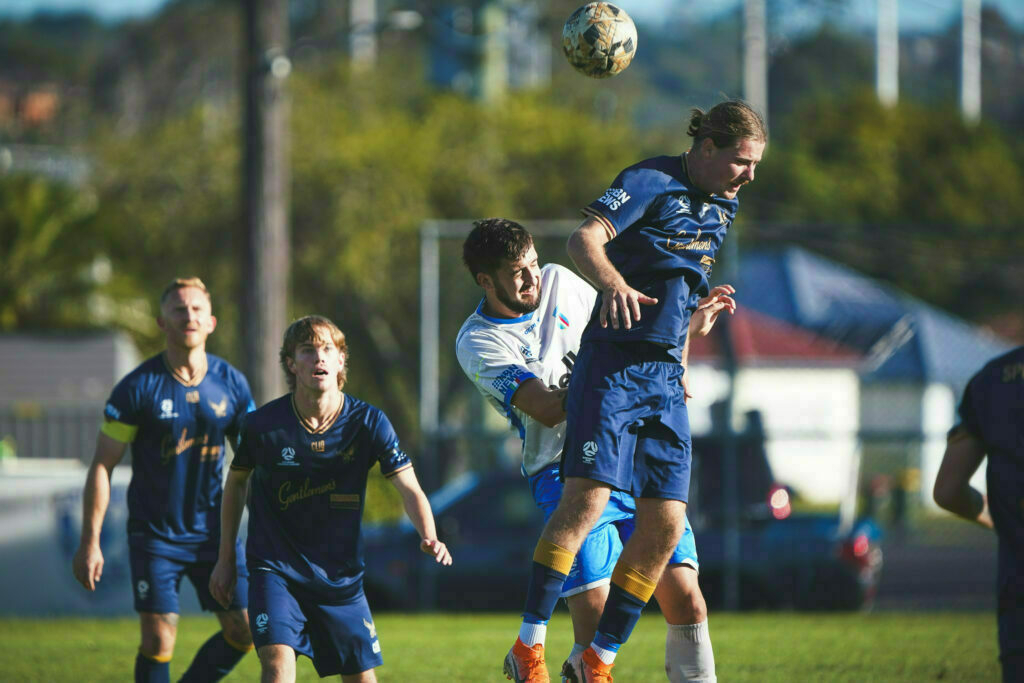 Two soccer players are competing to head the ball during a match, with other players observing in the background.