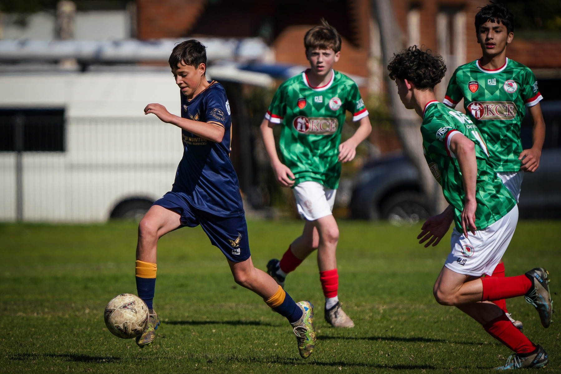 A young soccer player in a blue uniform dribbles the ball while being approached by opponents in green jerseys during a match.