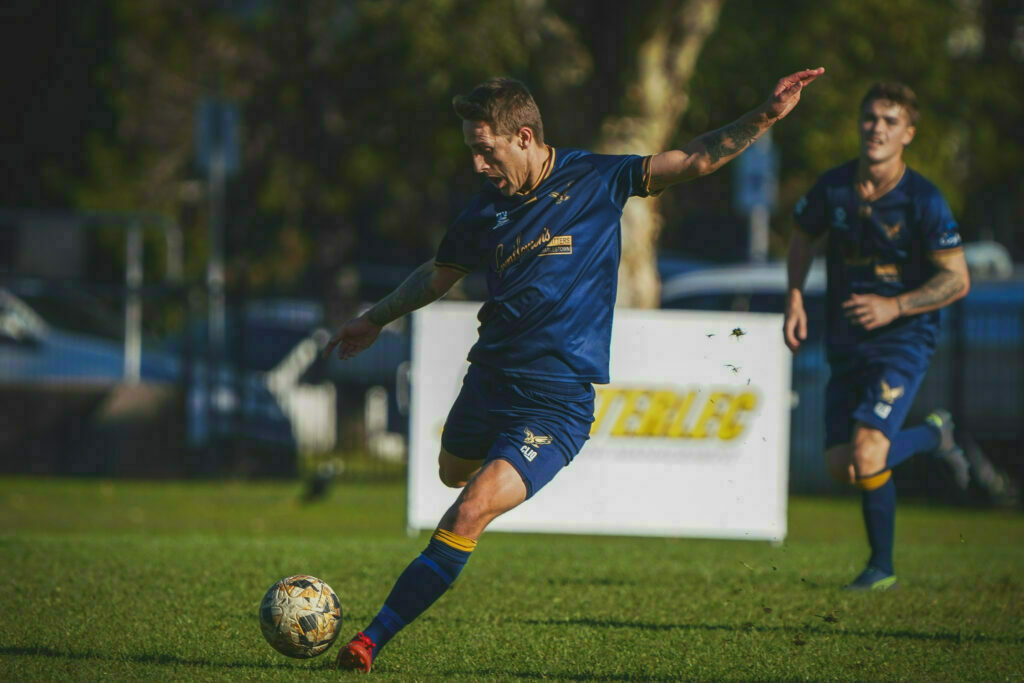 A soccer player in a blue uniform is about to kick a ball on a grassy field, with a teammate visible in the background.