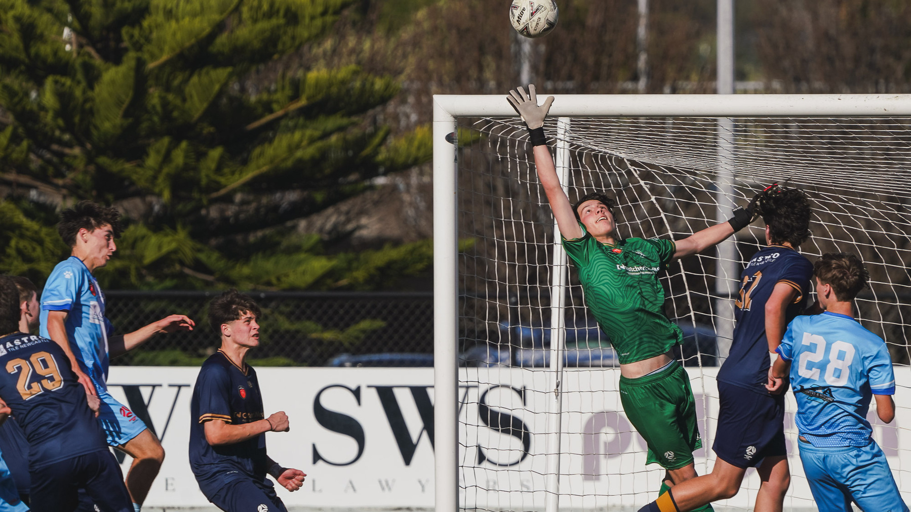 A goalkeeper in green leaps to block a high shot during a soccer match, surrounded by players in blue uniforms.