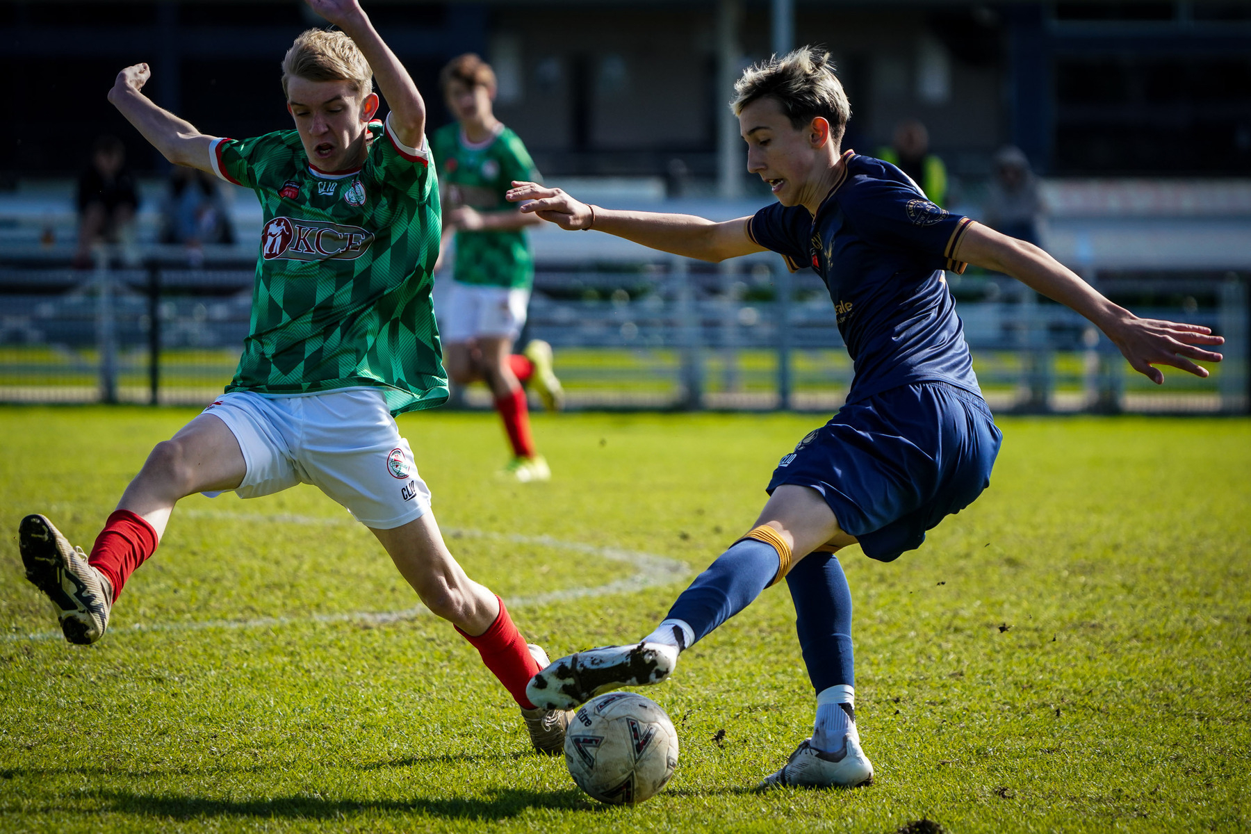 Two soccer players in action on the field, one in a green and red uniform and the other in blue, competing for control of the ball.