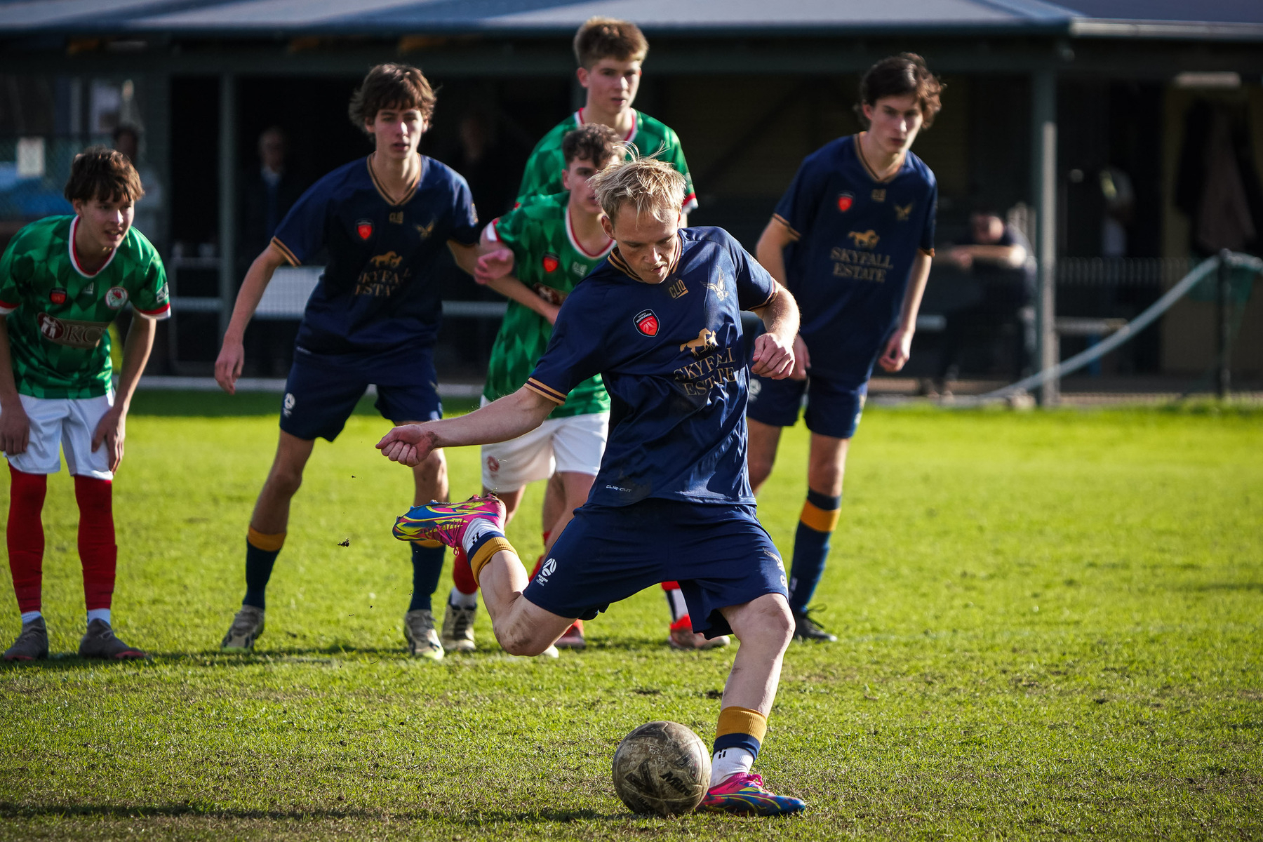 A soccer player in a blue jersey is taking a penalty kick while other players watch in the background.