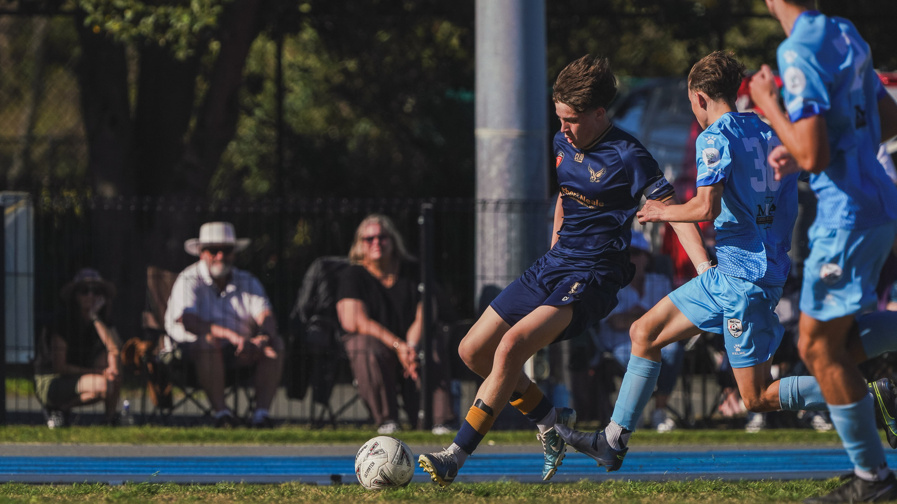 A soccer player in a blue uniform skillfully maneuvers the ball while pursued by opponents, with spectators watching from the sidelines.