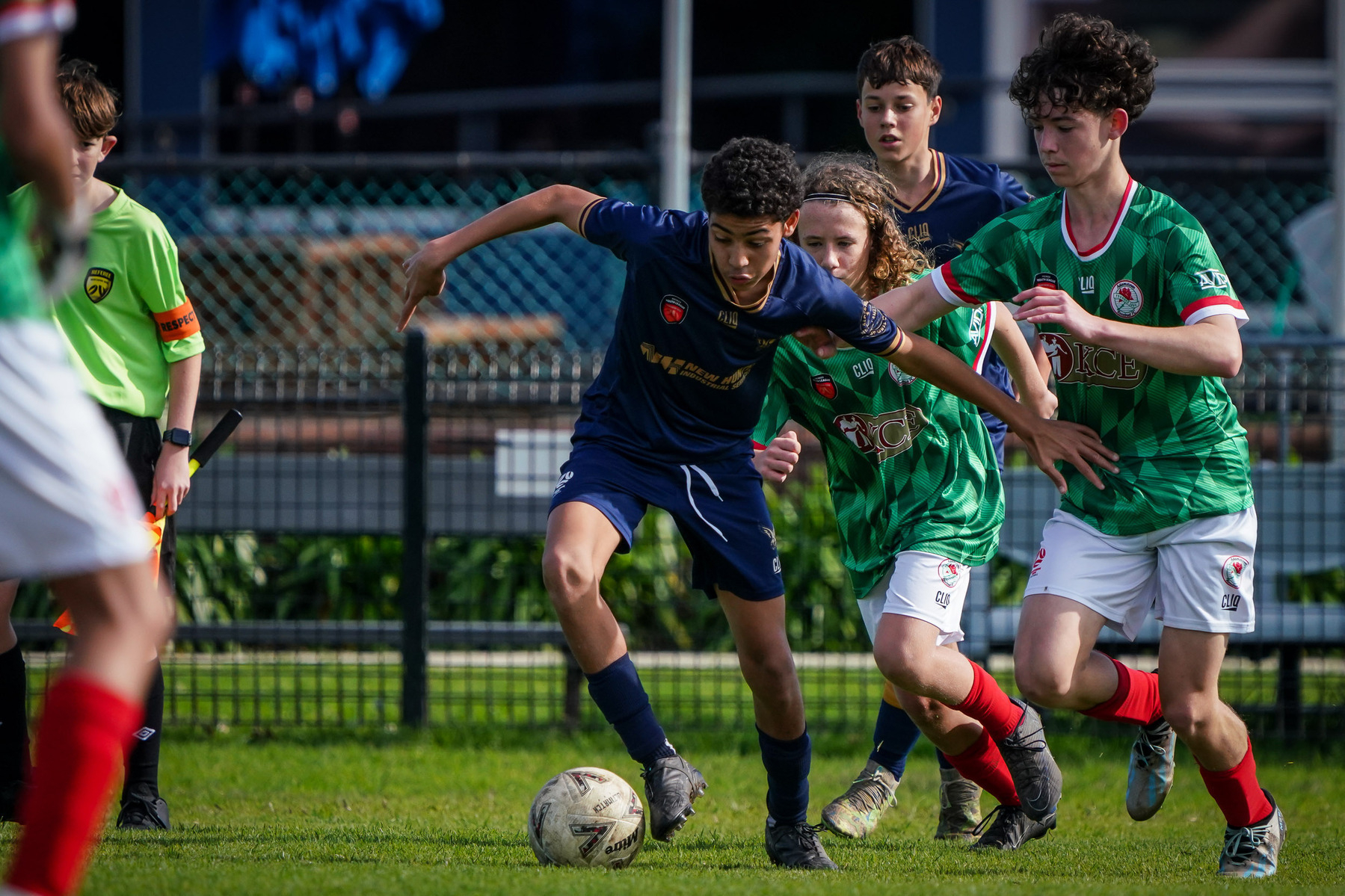 A group of young soccer players is energetically competing for the ball on a field, with a referee and spectators in the background.