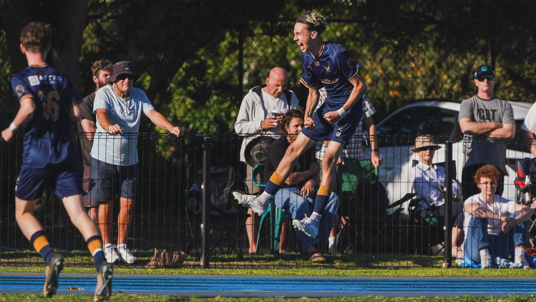 A soccer player in a blue jersey jumps in celebration while spectators watch from the sidelines.