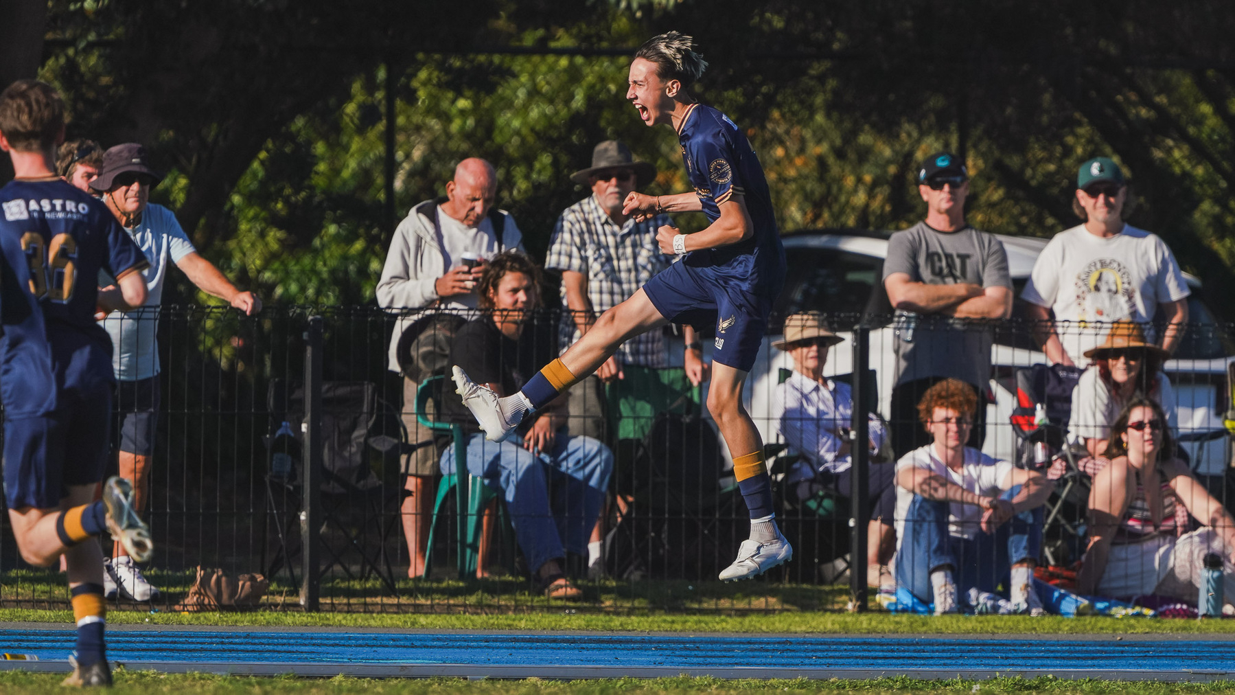 A soccer player leaps into the air in celebration while spectators watch from the sidelines.