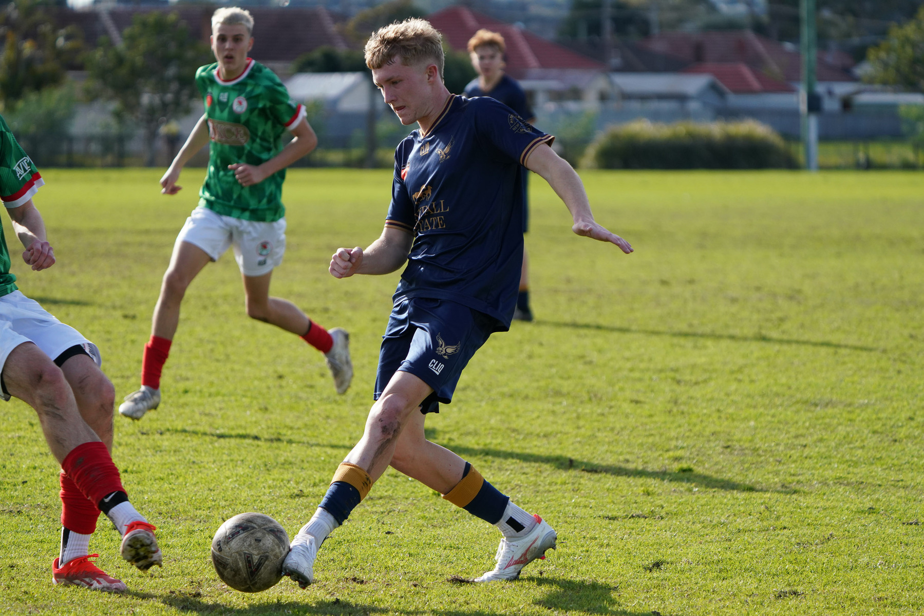 A soccer player in a navy blue uniform is poised to kick a ball during a match on a grassy field, with other players nearby.