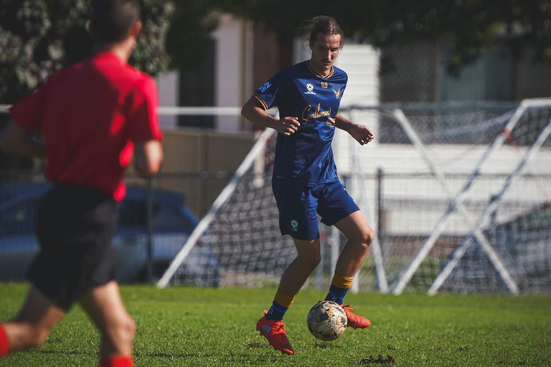 A soccer player in a blue uniform controls the ball on a grassy field with an official in red nearby.