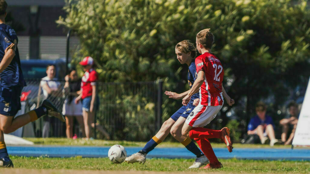 Two youth soccer players from opposing teams attempt to gain control of the ball during a match, with spectators watching in the background.