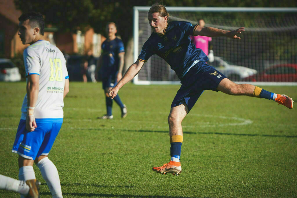 A soccer player in a blue jersey is in the action of kicking the ball during a match, with another player in a white jersey nearby.