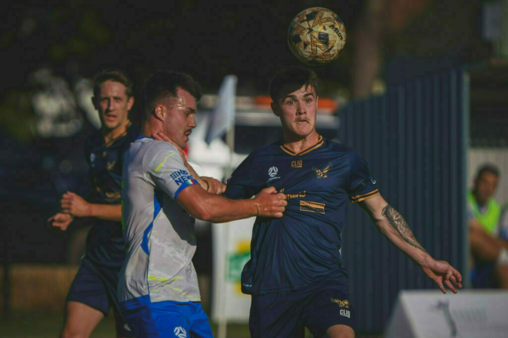 Two soccer players are competing intensely for the ball in mid-air during a match, with another player visible in the background.