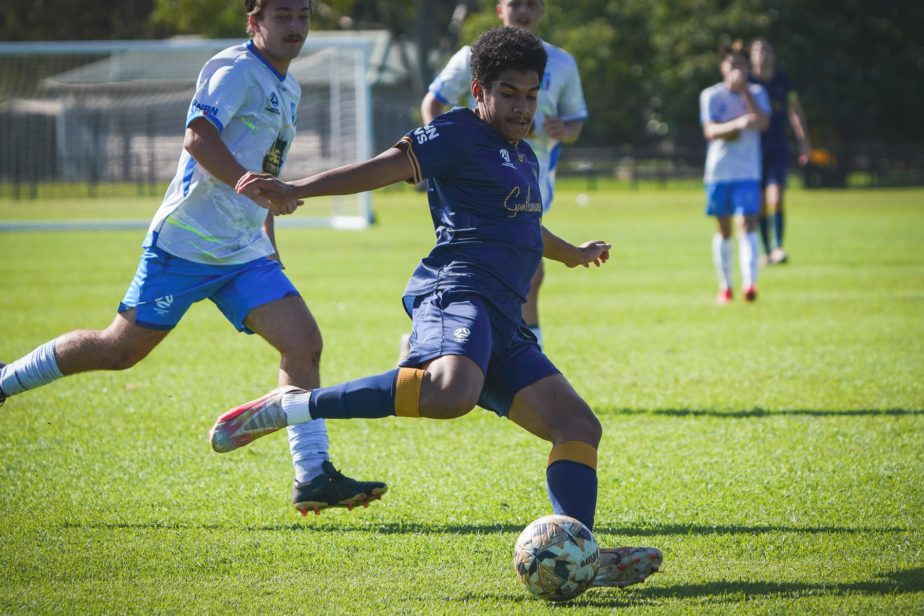 A soccer player in a blue uniform is attempting to kick the ball while being closely followed by an opponent in a white uniform during a match on a grassy field.