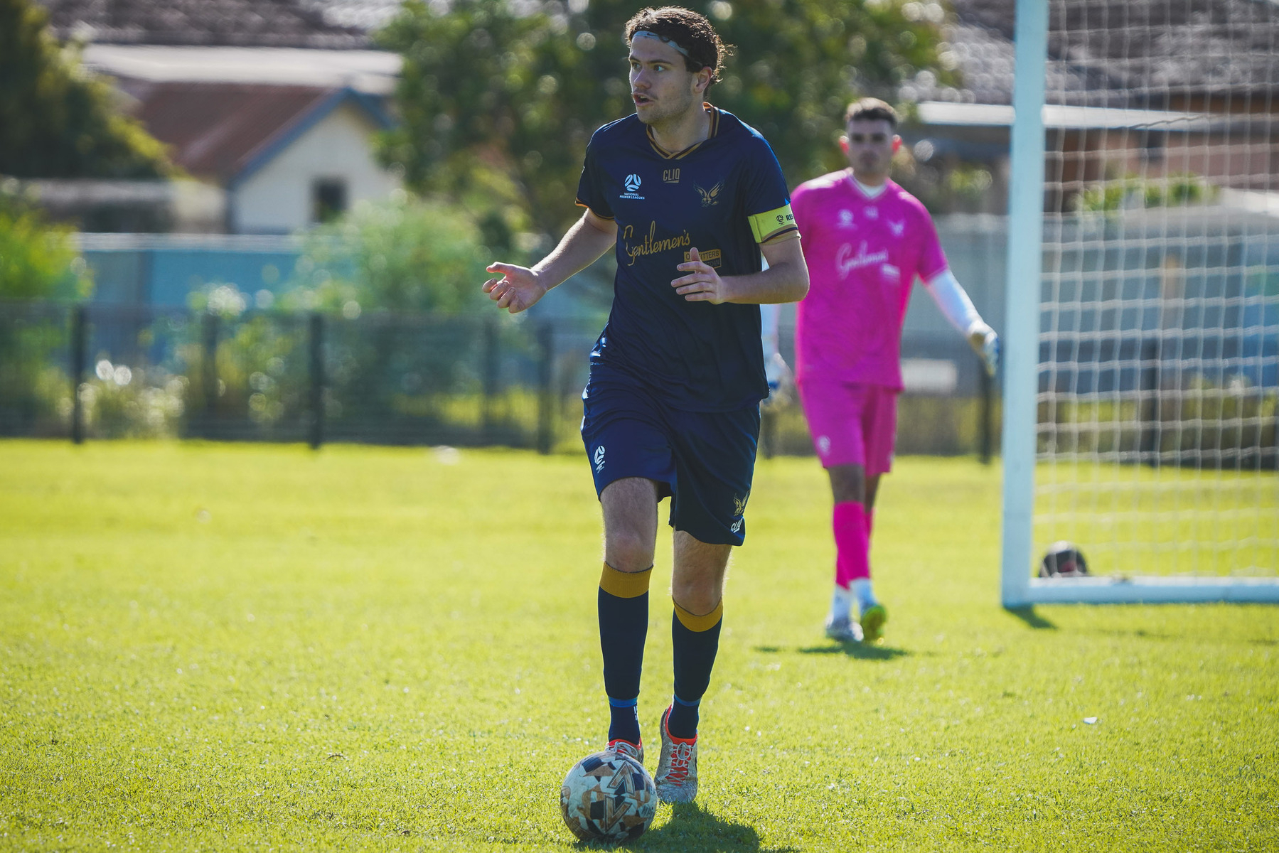 A soccer player in a blue uniform is dribbling the ball on a grassy field, with a goalkeeper in a pink uniform in the background near the goal.