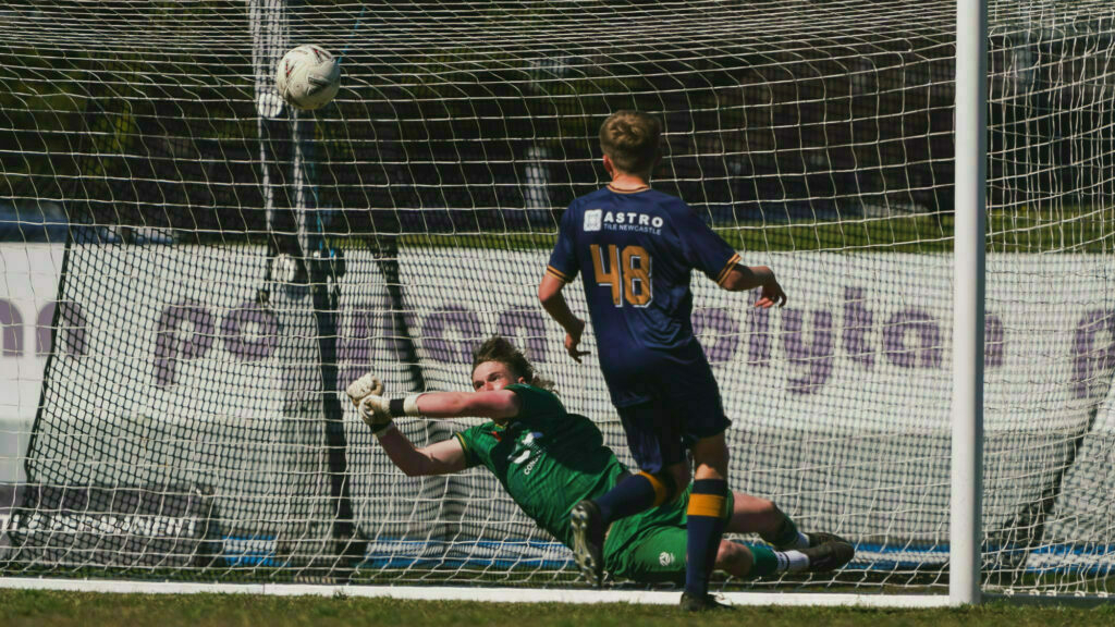 A soccer player in blue scores a goal against a goalkeeper in green, who is diving to make a save.