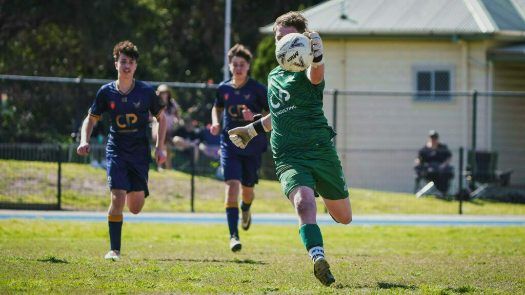 A soccer goalkeeper in green catches a ball while two players in blue run toward him on a grassy field.