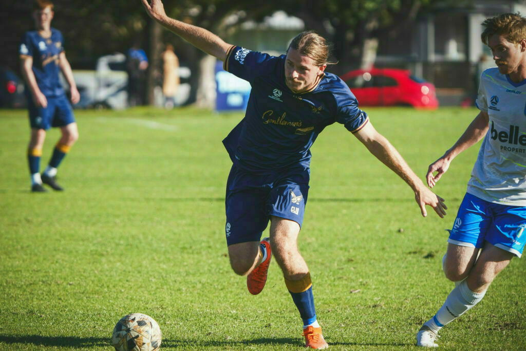 Two soccer players in blue uniforms are competing for the ball on a grassy field, with one player lunging forward.