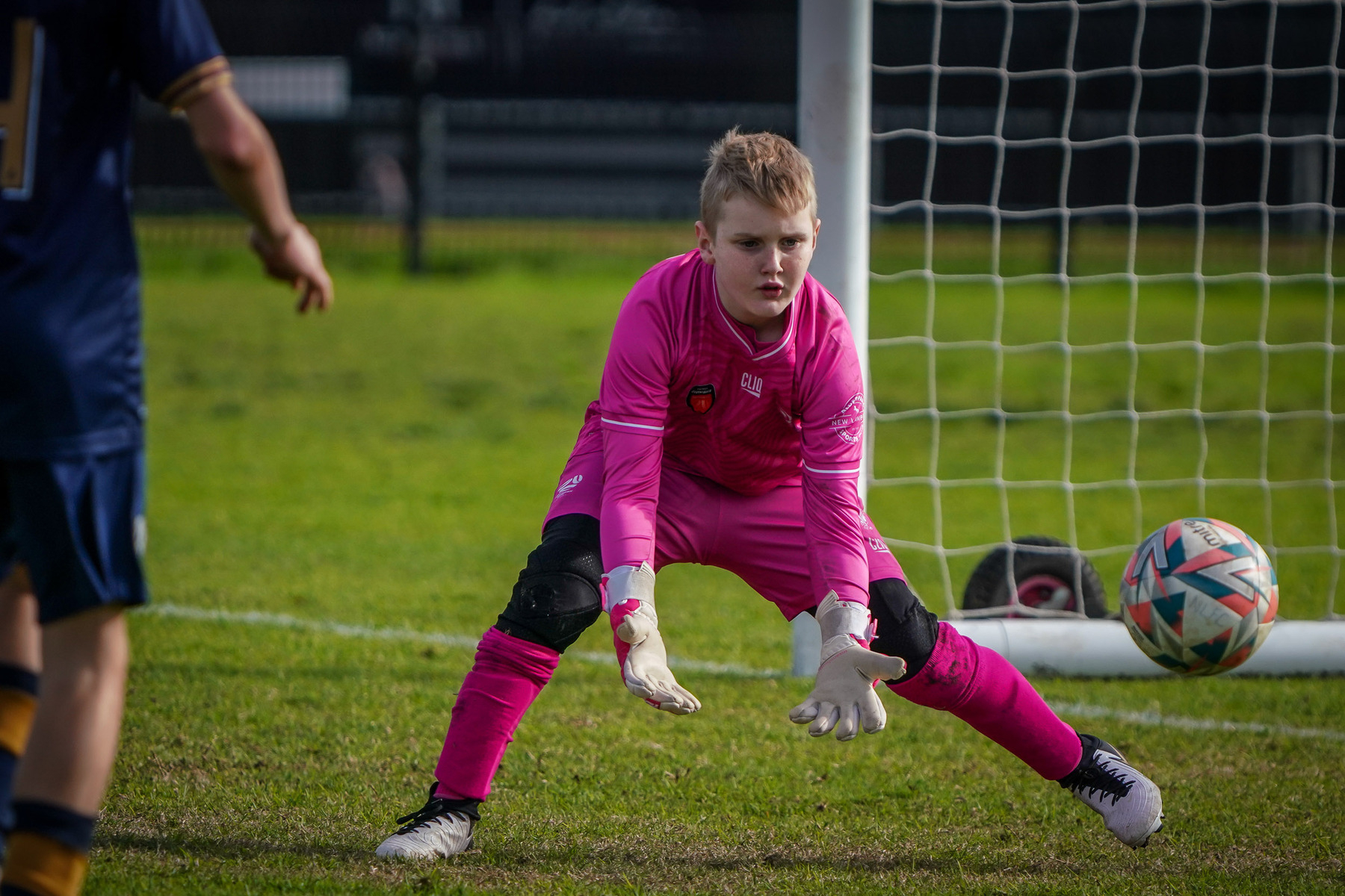 A young goalkeeper in a pink jersey is poised to catch an incoming soccer ball during a game.