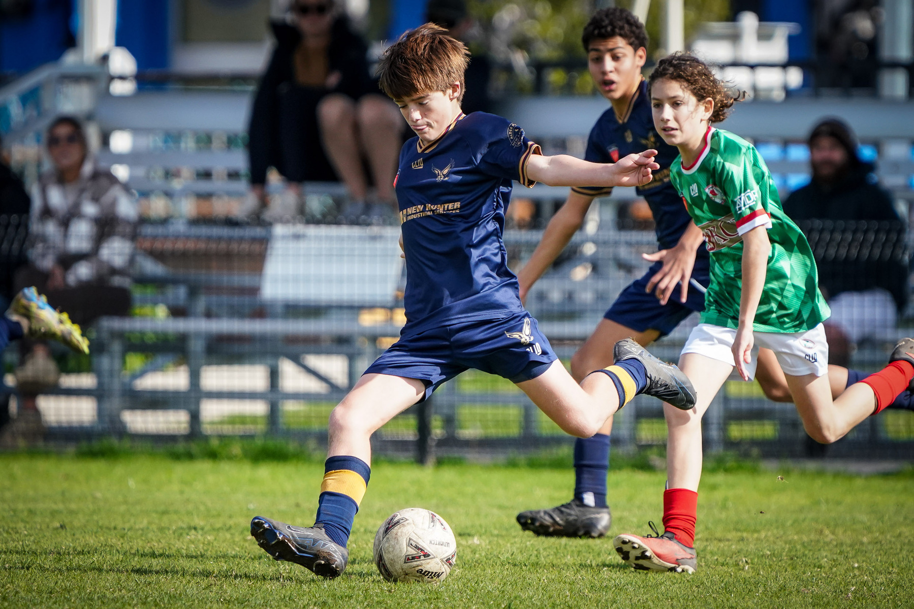 A young soccer player in a blue uniform is kicking a ball on a grassy field, with other players in the background.