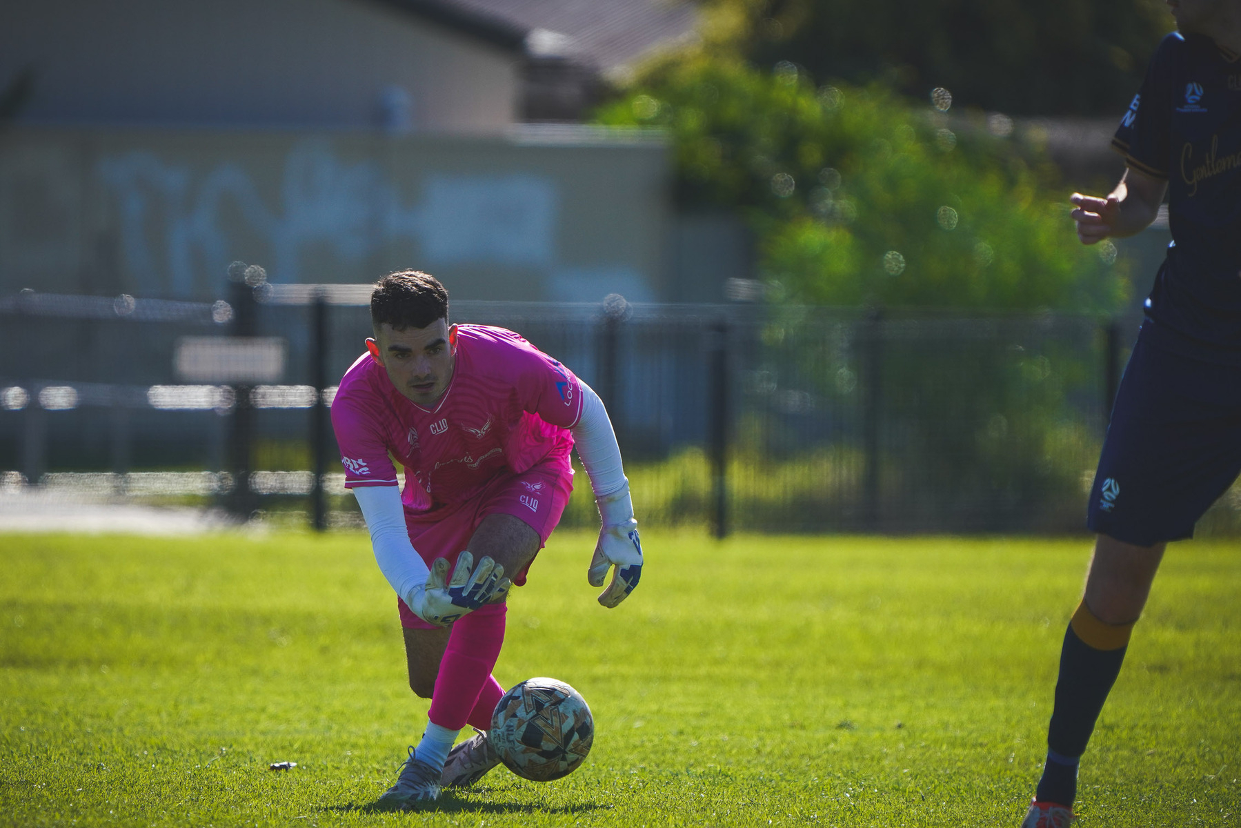 A soccer player wearing a pink uniform is bending down to pick up a ball on a grassy field.