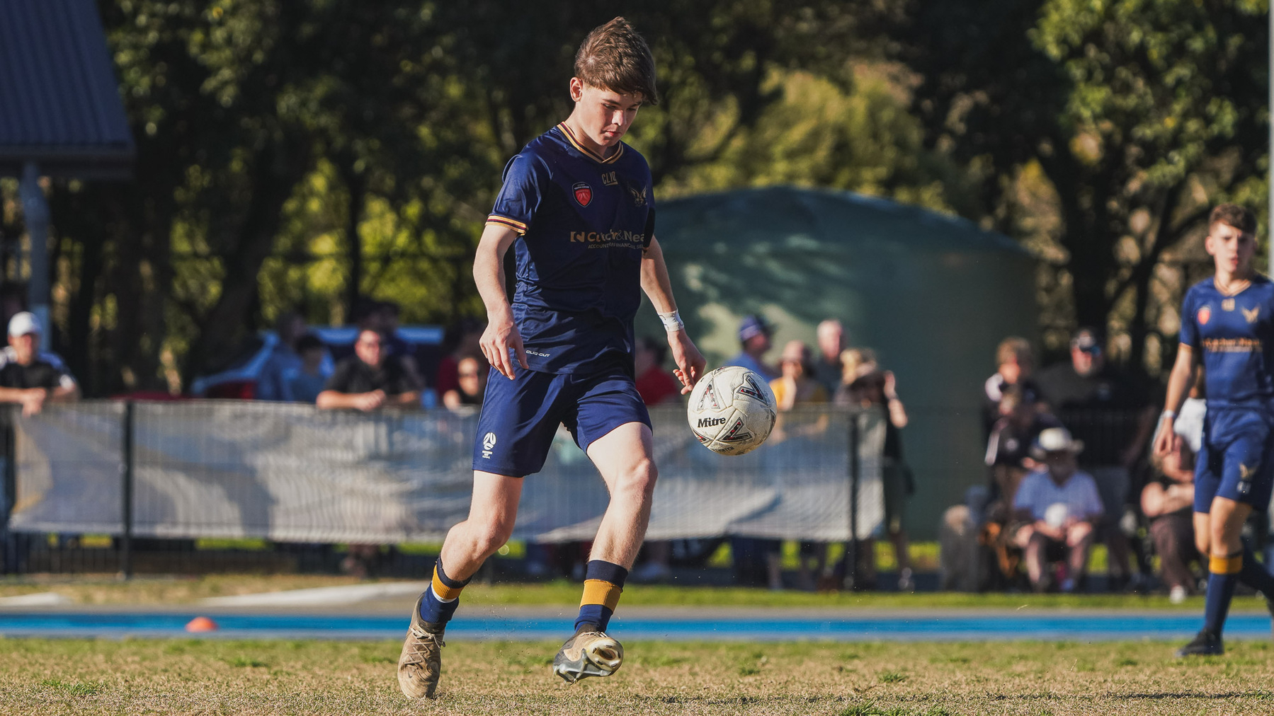 A soccer player in a blue uniform is controlling a ball on a grassy field during a match, with spectators in the background.