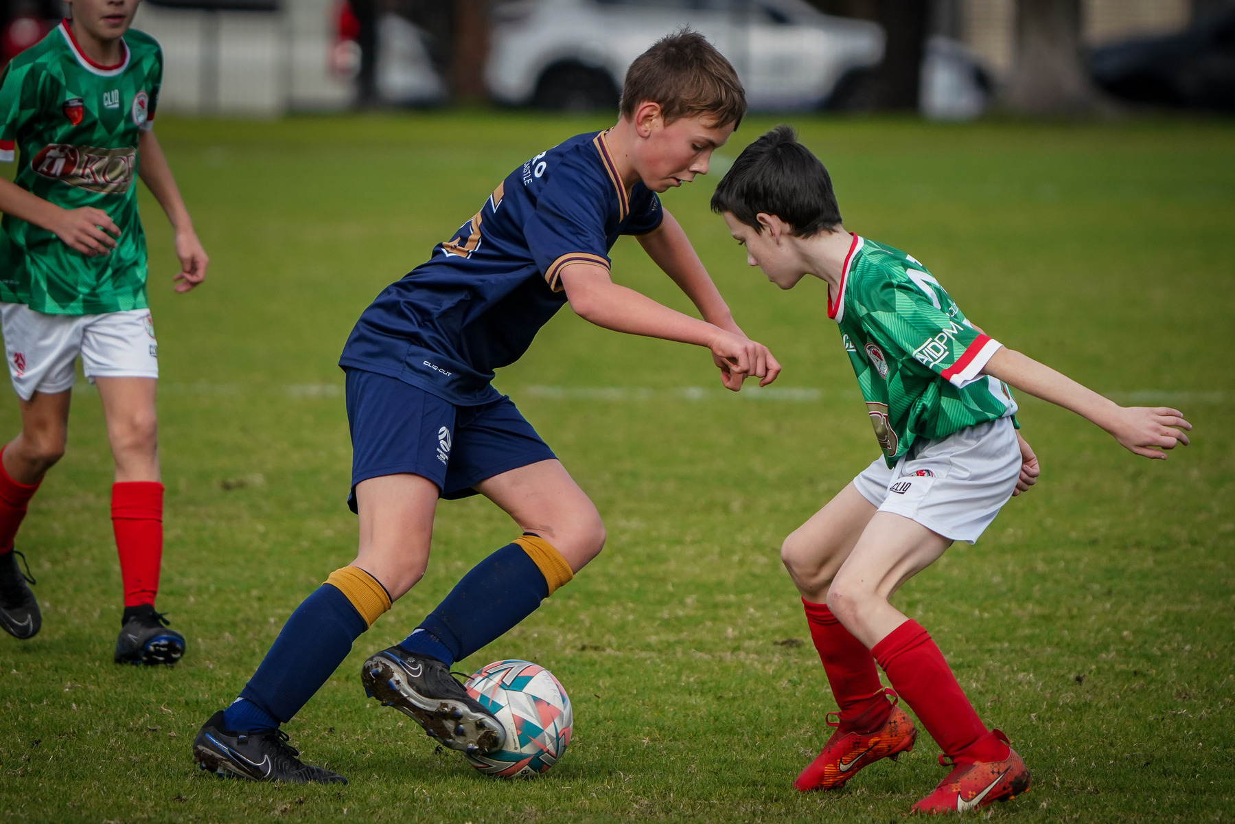 Two youth soccer players are competing for the ball on a grassy field, surrounded by teammates.